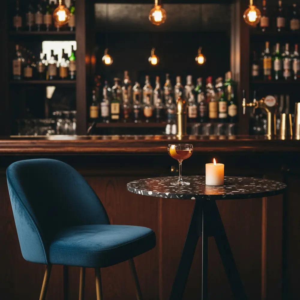 Round black table with recycled plastic terrazzo top in a moody cocktail bar, paired with a blue velvet chair and lit candle