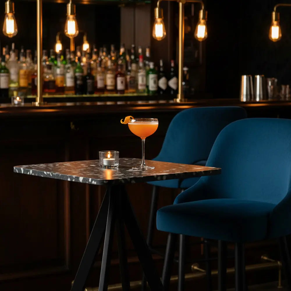 Square black HDPE table with speckled panel top in an upscale bar, styled with blue velvet chairs and a garnished cocktail