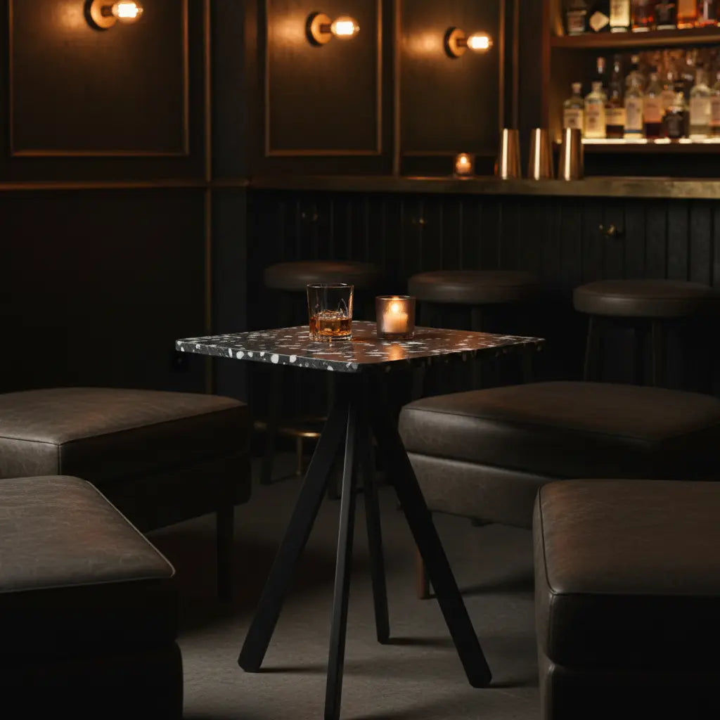 Square black polymer table made from recycled plastic panels in a dark lounge, with a whiskey glass, candle, and leather ottomans