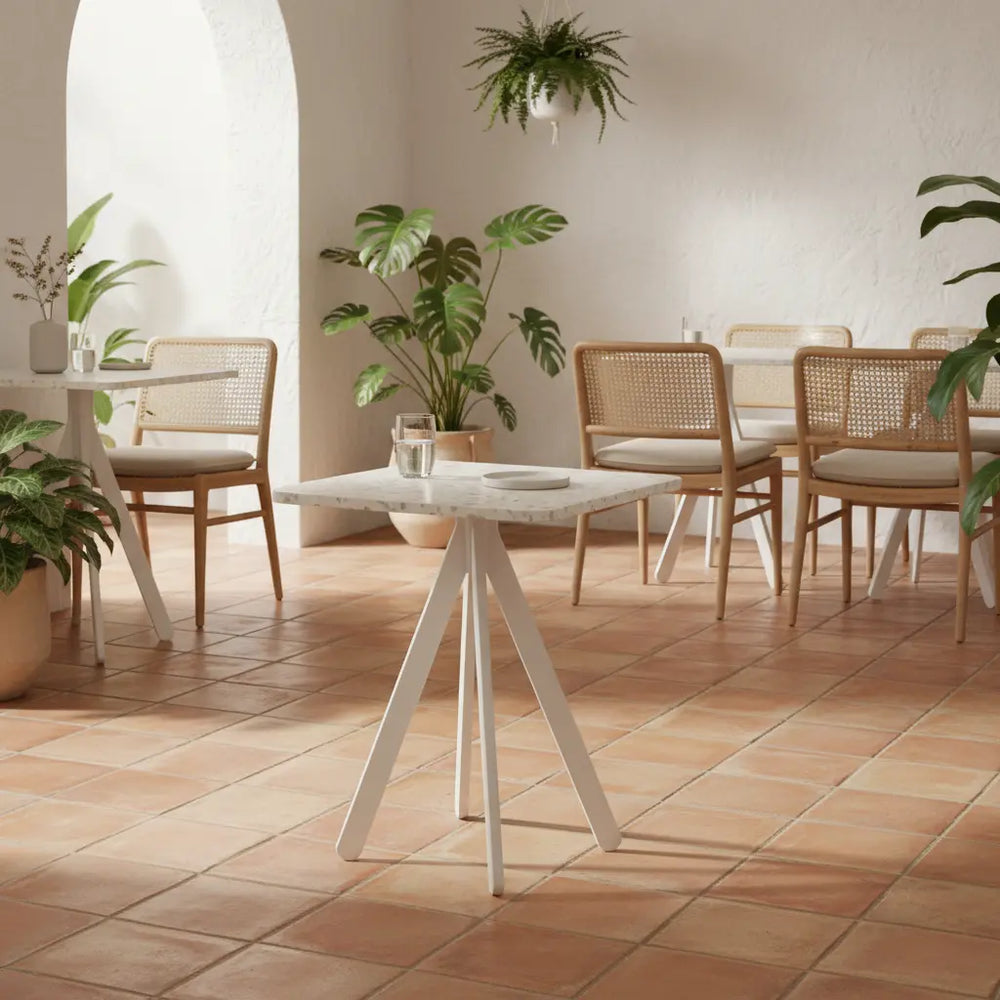 Square white polymer table with recycled panel top in a HoReCa space with terracotta flooring and rattan chairs